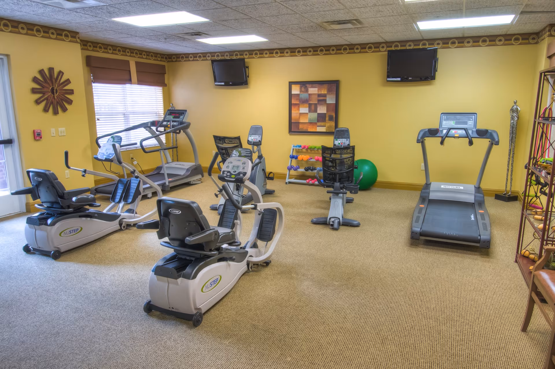 A fitness room with exercise equipment including two recumbent bikes, two treadmills, and two upright bikes. The room has yellow walls, a beige carpet, two wall-mounted televisions, a rack of colorful dumbbells, and a large green exercise ball. There is a window with brown blinds and a decorative wall clock.