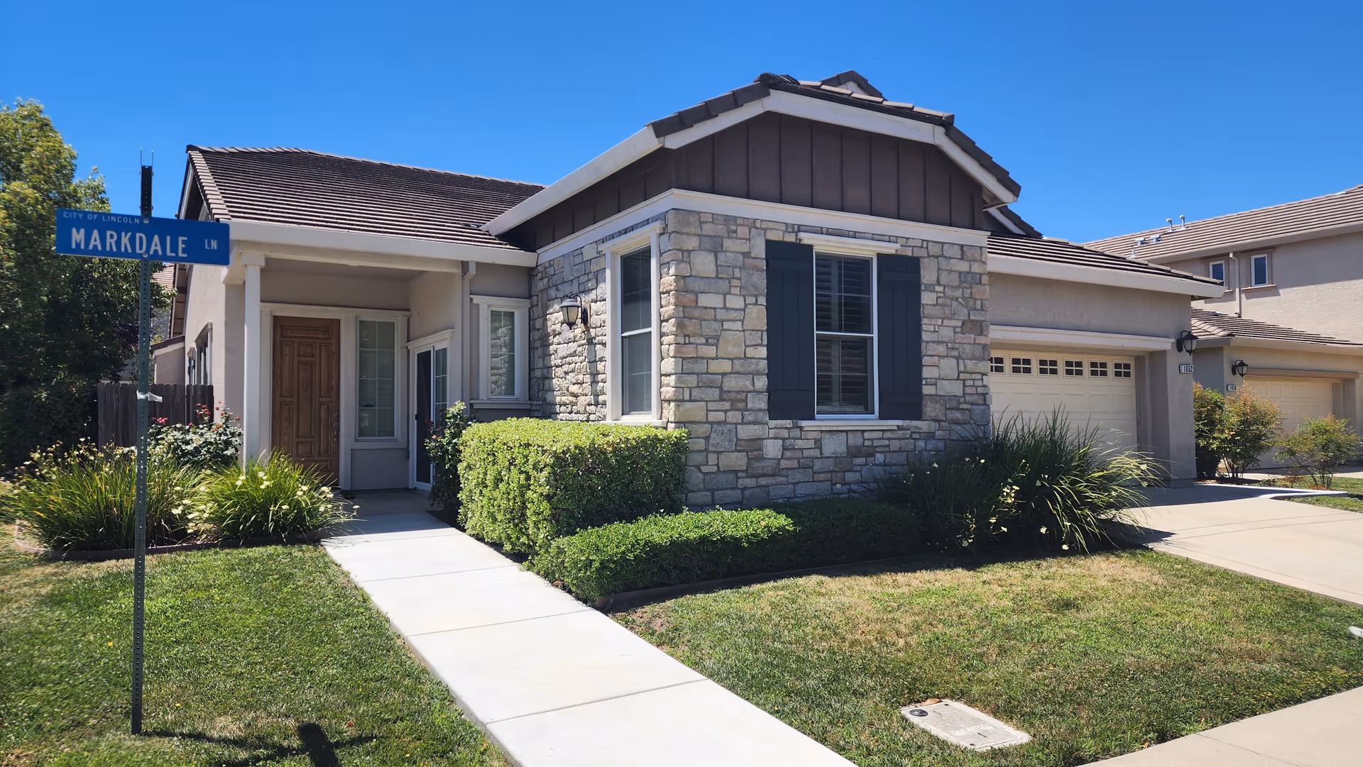 Exterior view of a single-story residential building with stone and stucco facade, a wooden front door, and a garage. The building is surrounded by a well-maintained lawn, bushes, and a sidewalk leading to the entrance. A street sign labeled 'Markdale Ln' is visible on the left side under a clear blue sky.