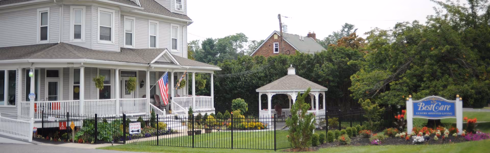 Exterior view of a large, multi-story house with a wraparound porch, white railings, and an American flag displayed near the entrance. The house is surrounded by a black metal fence and well-maintained landscaping with flowers and shrubs. A white gazebo is visible in the fenced yard, and a blue sign reading 'BestCare Luxury Assisted Living' is positioned near the entrance to the property.