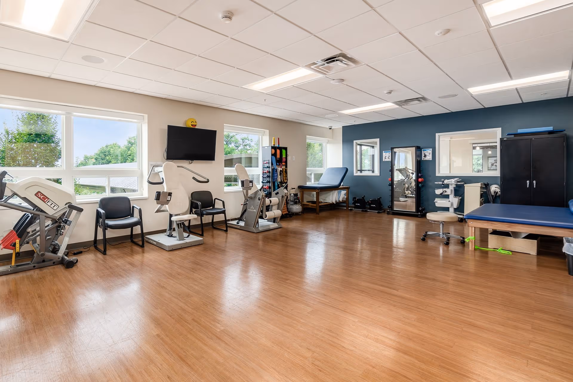 Spacious physical therapy/exercise room with rehabilitation equipment, treatment tables, chairs, and large windows.