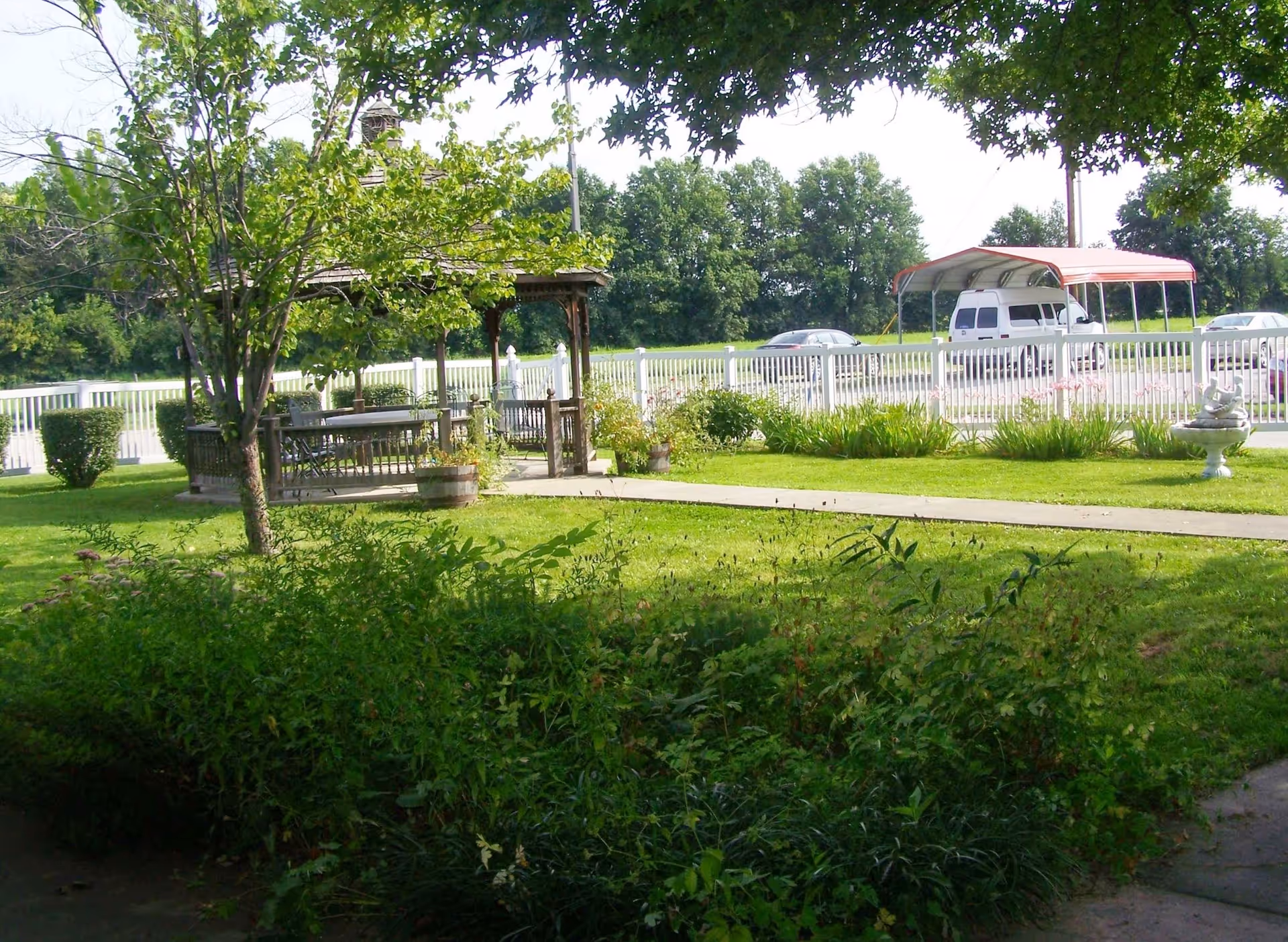 Outdoor lawn and garden with a wooden gazebo and seating, a white fence, and parked vehicles in the background.