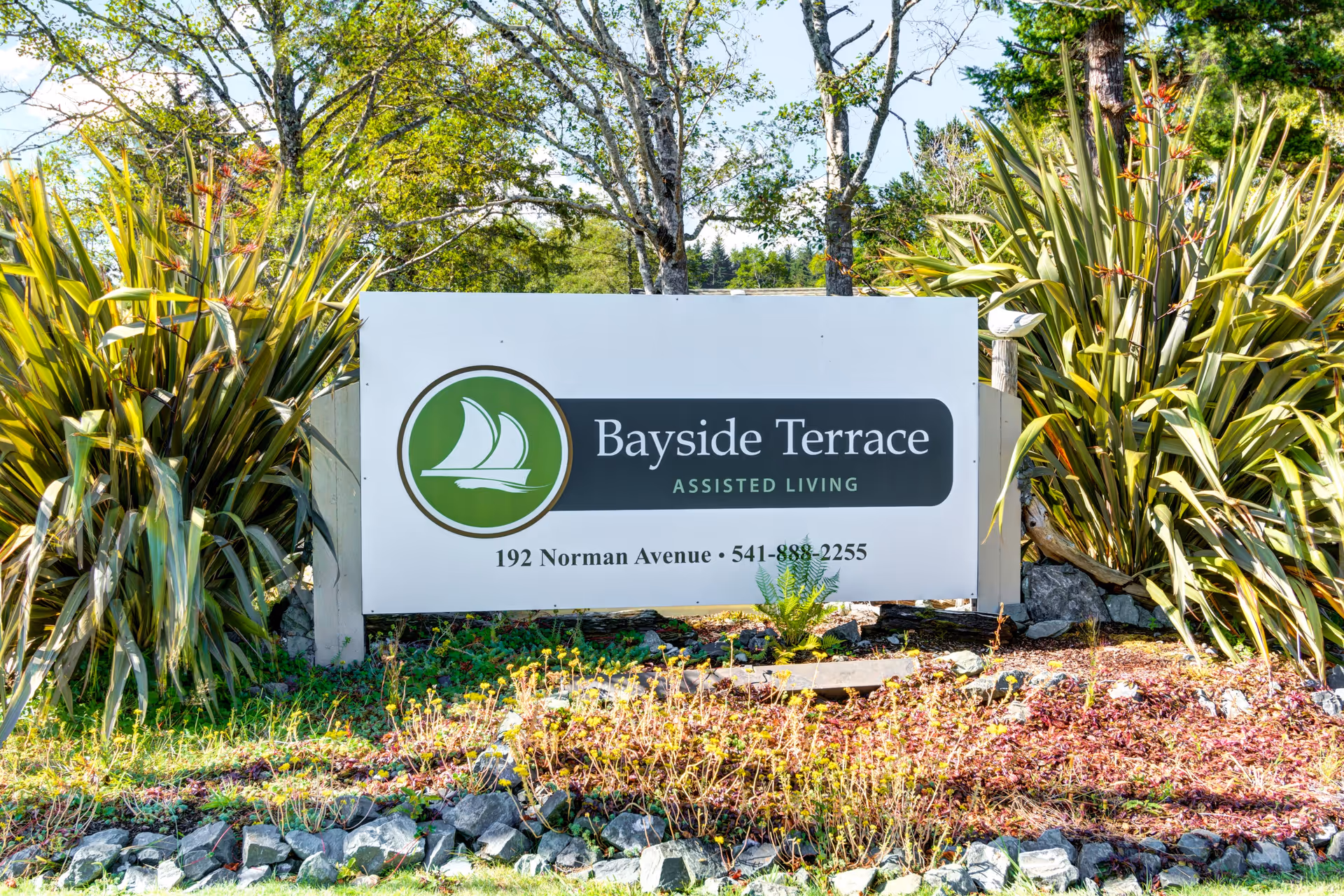 Outdoor sign for Bayside Terrace Assisted Living located at 192 Norman Avenue with a phone number, surrounded by plants and trees in a landscaped area.