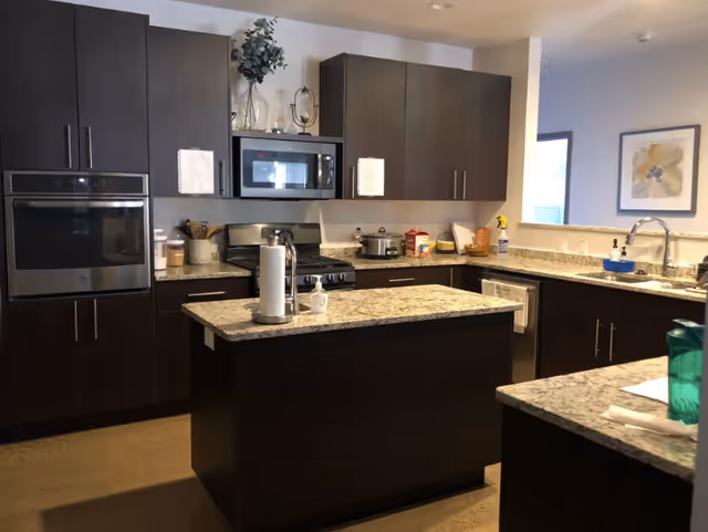 Modern kitchen with dark wood cabinets, granite countertops, a central island, built-in oven, microwave, stove, and various kitchen items such as a paper towel holder, hand sanitizer, and small appliances.