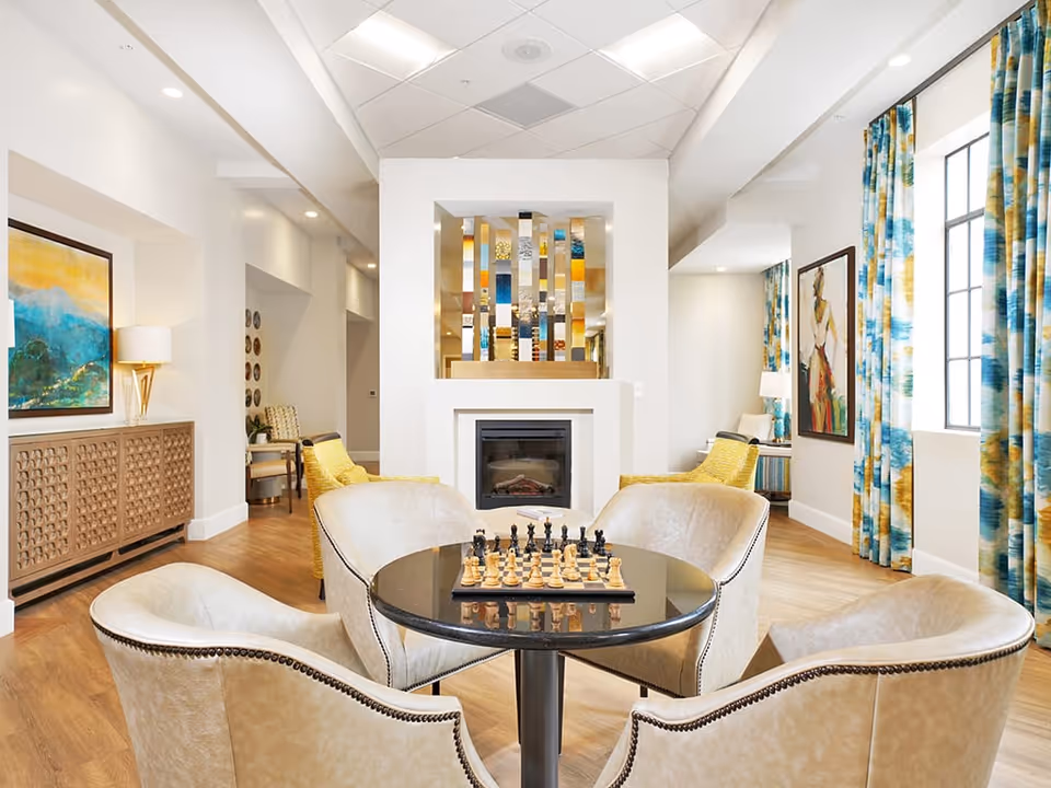 A bright and inviting living room area with a round black table in the center holding a chessboard. Four beige upholstered chairs surround the table. Behind the table is a modern fireplace with a decorative mirrored panel above it. The room features wooden flooring, colorful blue and yellow curtains on the windows, and framed artwork on the walls. Additional seating with yellow chairs and side tables with lamps are visible in the background.