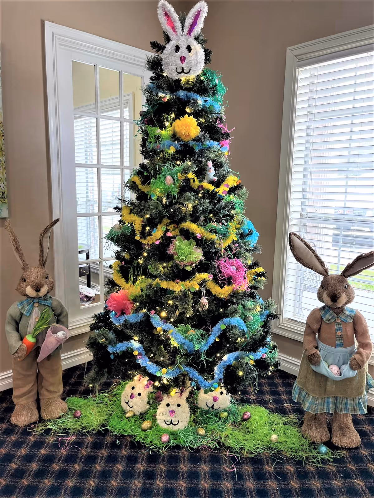 A decorated Christmas tree with colorful garlands and bunny face ornaments, topped with a bunny head. Two large stuffed bunny figures stand on either side of the tree, one holding a carrot and the other holding Easter eggs in an apron. The scene is set indoors near windows with blinds and a glass door.