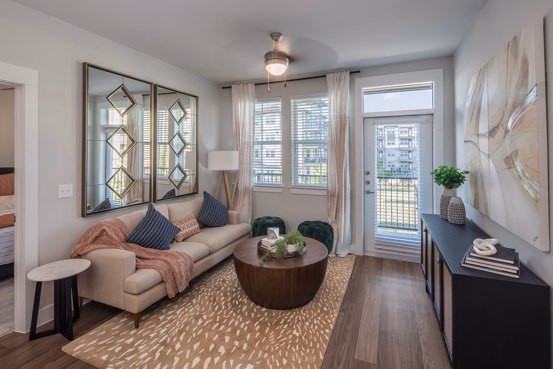 Bright furnished living room with a sofa, round coffee table, decorative mirrors, and a glass door and windows leading outside.