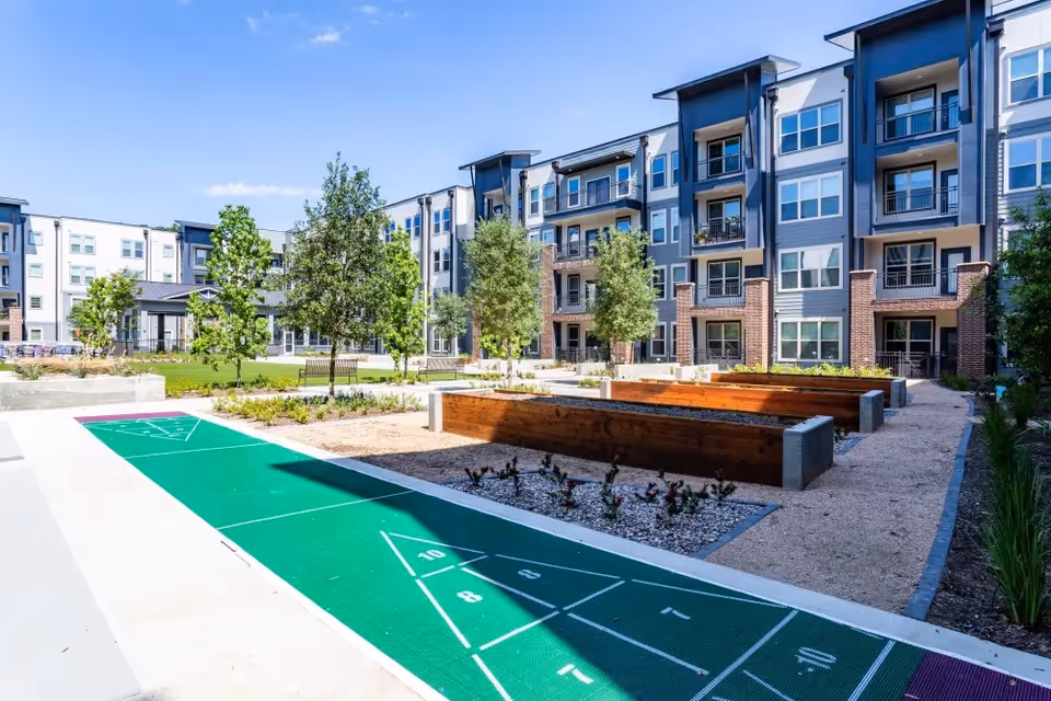 Sunny courtyard of a modern multi-story senior living building with a shuffleboard court, raised planter beds, trees, and balconies.