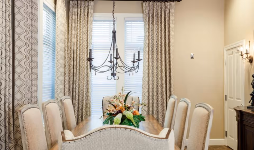 Elegant dining room with a wooden table, upholstered chairs, a chandelier, floral centerpiece, and patterned curtains over tall windows.