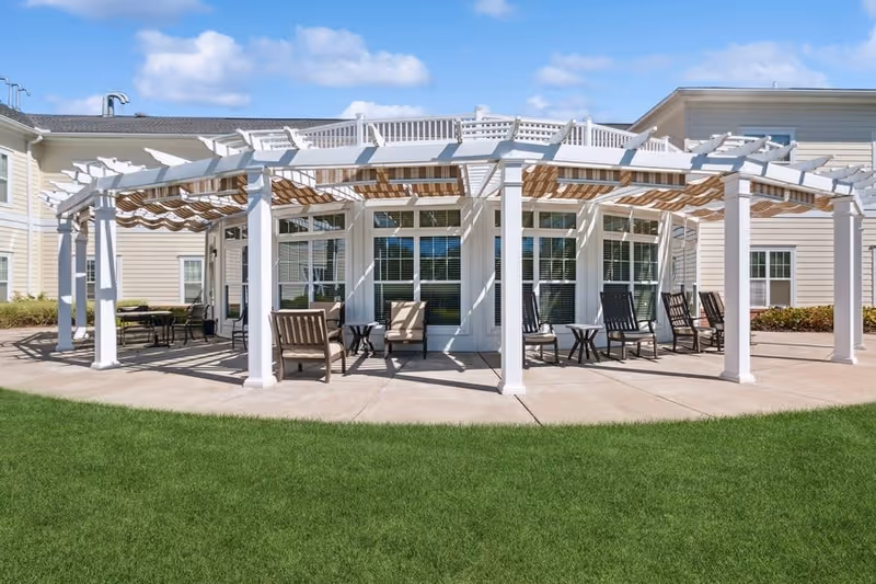 Outdoor patio area at The Bridges at Warwick featuring a white pergola with striped fabric shades, several chairs and small tables arranged underneath, adjacent to a beige building with large windows and a well-maintained green lawn in the foreground.