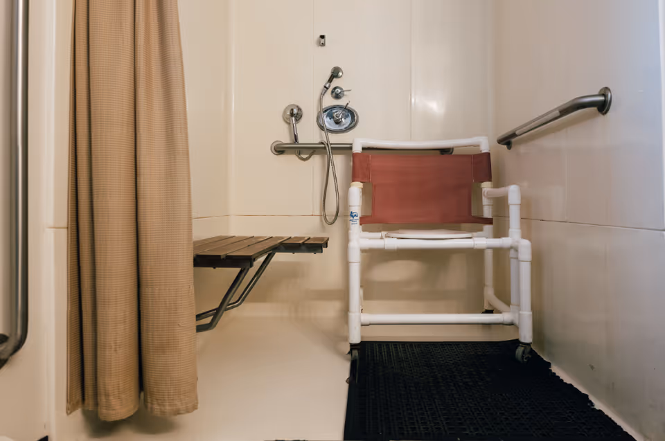 Accessible shower area with a beige curtain, a foldable wooden shower seat attached to the wall, a handheld showerhead, grab bars on the walls, and a white PVC frame shower chair with a maroon fabric backrest and seat on a black rubber mat.