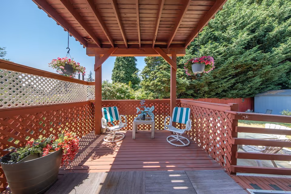 Covered wooden patio with two striped chairs, a small table, hanging flower baskets, and lattice railings overlooking greenery.