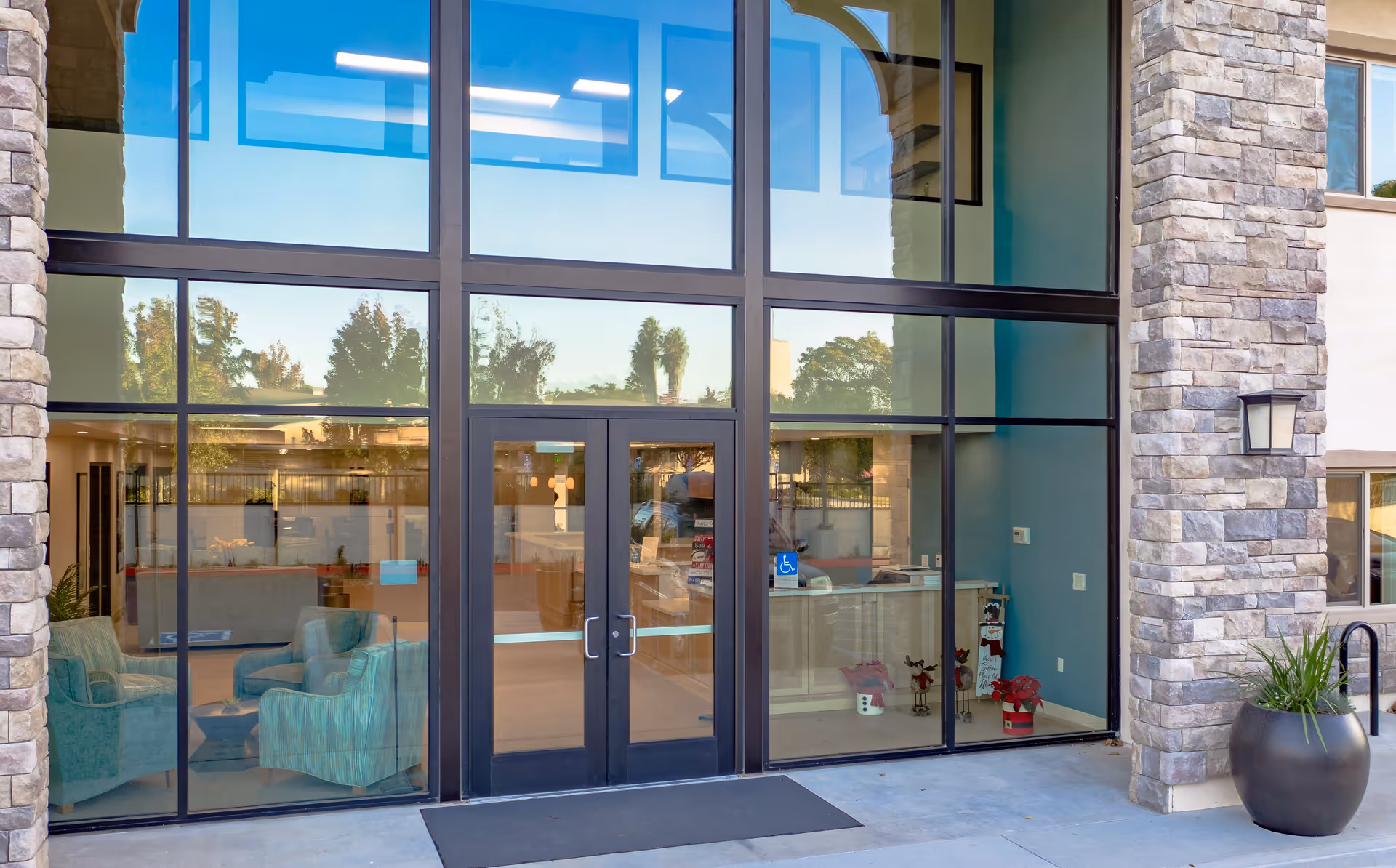 Glass entrance doors to a building with large windows on either side, showing a seating area with teal armchairs and a reception desk inside. The exterior features stone pillars and a large planter with greenery.