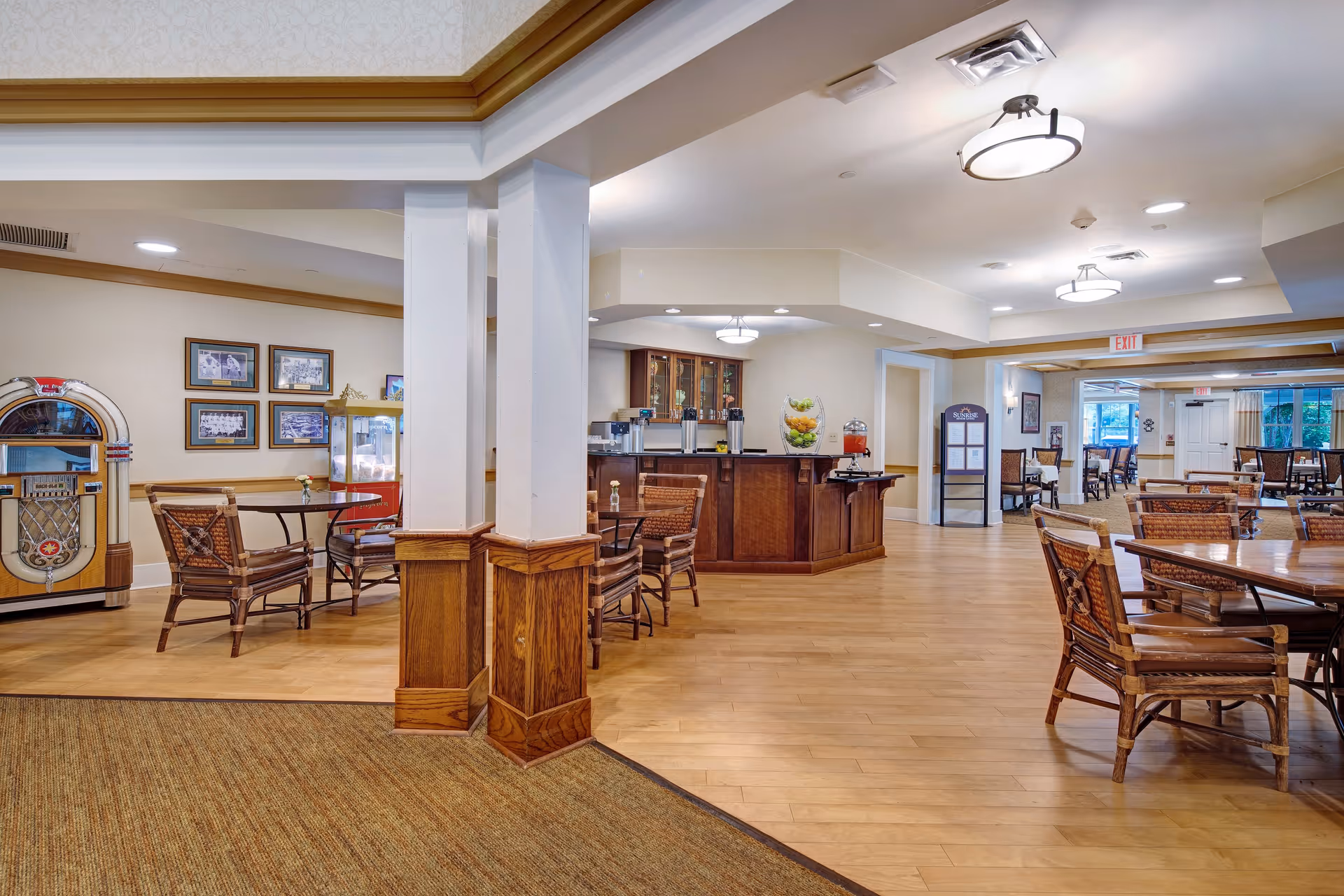 Interior view of a senior living facility dining area with wooden floors and furniture. There are multiple tables and chairs arranged for seating, a jukebox on the left side, framed pictures on the wall, and a counter with coffee dispensers and a fruit display. The ceiling has recessed lighting and modern light fixtures.