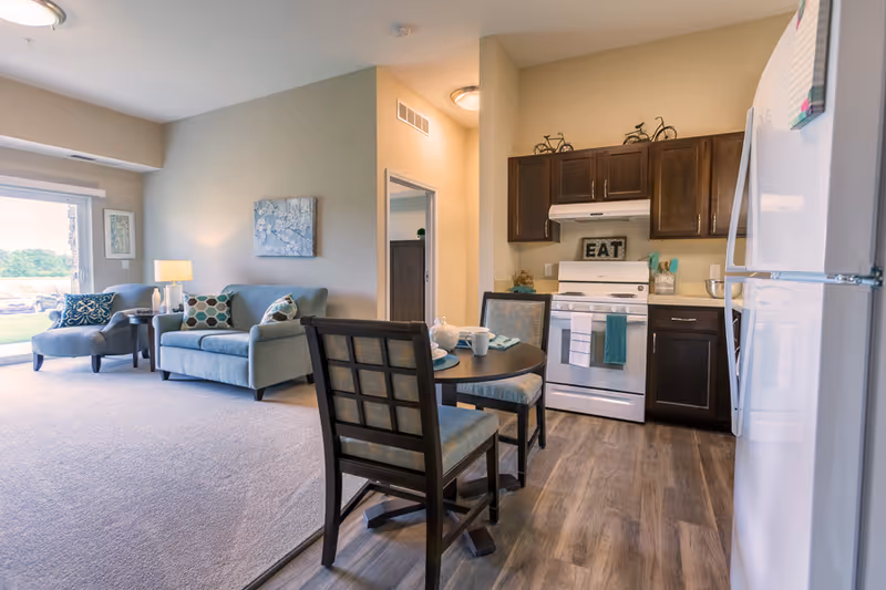 A bright and cozy living space featuring a small kitchen with dark wood cabinets, a white stove, and a white refrigerator. In front of the kitchen is a round dining table with two chairs set with plates and cups. Adjacent to the kitchen is a living room area with a light blue sofa, a matching armchair, a side table with a lamp, and wall art. A large window lets in natural light and offers a view of greenery outside.