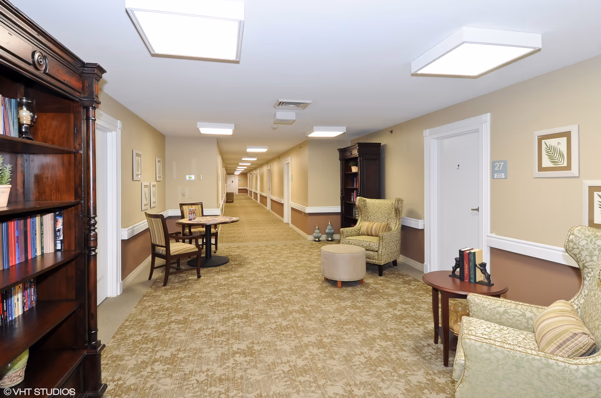 Well-lit senior living hallway with carpeted floor, seating areas, bookshelves and doors to resident rooms.
