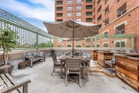 Outdoor patio area with a large umbrella over a table surrounded by chairs, wooden benches with cushions, potted plants, and a tall brick building in the background under a partly cloudy sky.