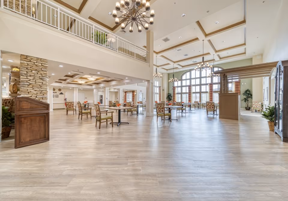 Spacious and well-lit dining area in a senior living facility with multiple tables and chairs arranged neatly. The room features large windows allowing natural light to fill the space, high ceilings with decorative chandeliers, and a second-floor balcony overlooking the area. The decor includes stone pillars, plants, and patterned upholstery on the chairs.