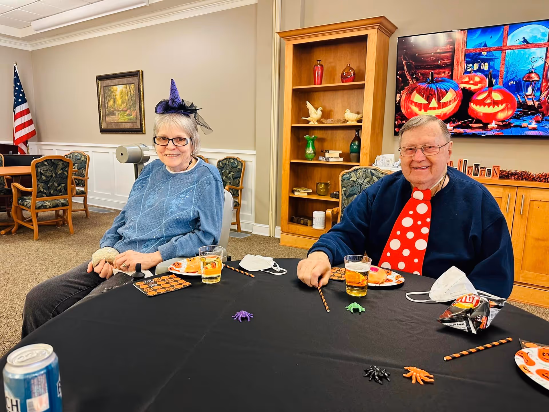 Two elderly people sitting at a table decorated for Halloween with small plastic spiders and Halloween-themed napkins. The woman on the left is wearing a small purple witch hat and a blue sweater, while the man on the right is wearing a large red tie with white polka dots and a dark sweater. Behind them is a wooden bookshelf with decorative items and a TV screen displaying a Halloween scene with carved pumpkins. An American flag and framed painting are visible in the background.