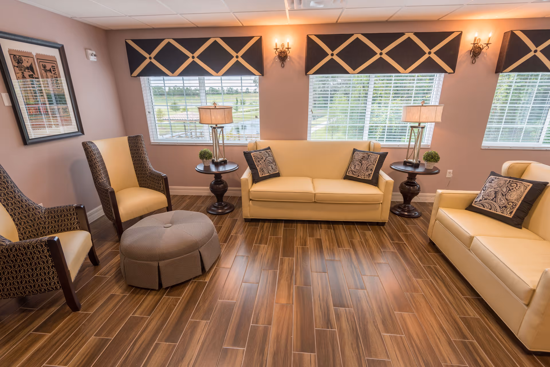 A cozy living room with two beige sofas and two patterned armchairs arranged around a round ottoman on a wooden floor. The room has three large windows with blinds and decorative valances, two table lamps on dark wooden side tables, and framed artwork on the wall.