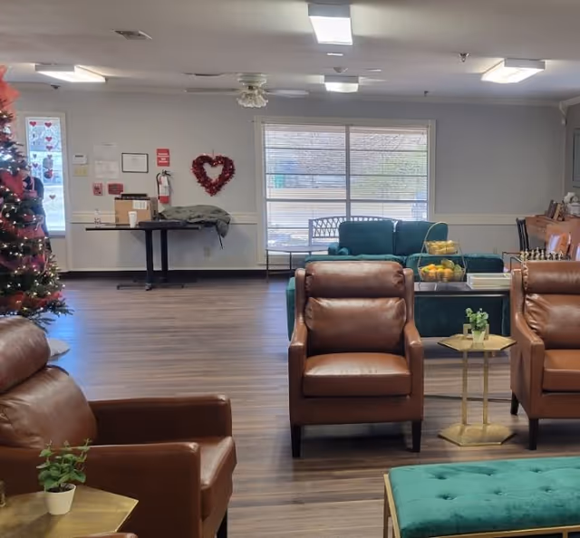 A spacious assisted living facility common area with brown leather armchairs, a green upholstered bench, and a green sofa near a large window. There is a decorated Christmas tree on the left, a small table with a potted plant, and a table with fruit baskets and books. The room has wood flooring and fluorescent ceiling lights.