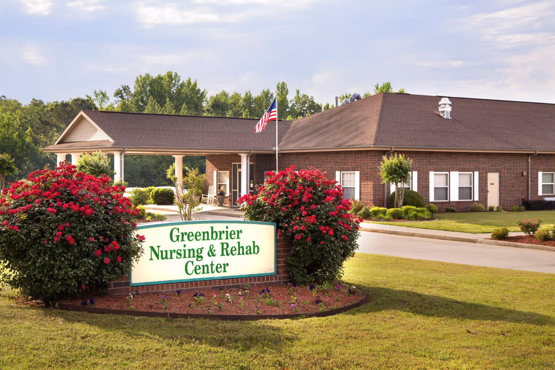Exterior view of Greenbrier Nursing & Rehab Center showing a single-story brick building with a covered entrance, an American flag on a pole, and well-maintained landscaping including green grass and red flowering bushes around a sign with the facility's name.