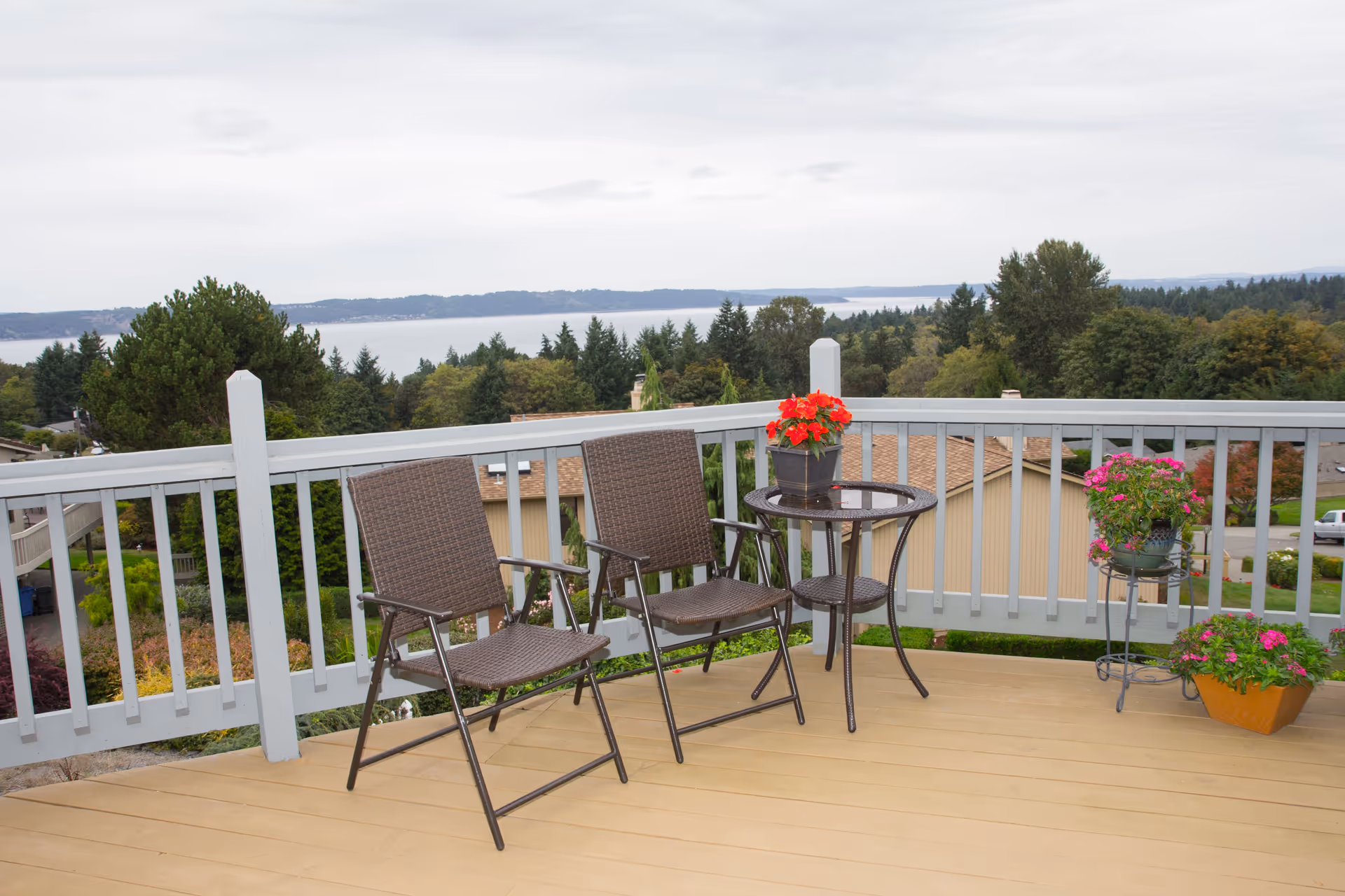 Outdoor deck area with two brown wicker chairs and a small round glass-top table holding a red flower pot. The deck has a light gray railing and overlooks a scenic view of trees, rooftops, and a body of water in the distance under a cloudy sky. Additional flower pots with pink flowers are placed on the deck floor and a metal plant stand.