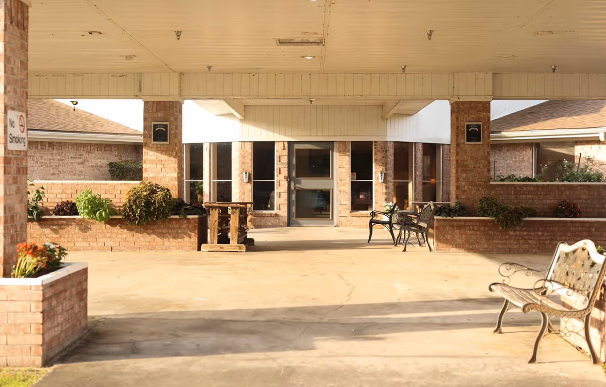 Covered outdoor seating area with brick walls and planters, featuring metal benches and a wooden table. The area is part of a building entrance with glass doors and windows, and a 'No Smoking' sign is visible on a brick pillar.