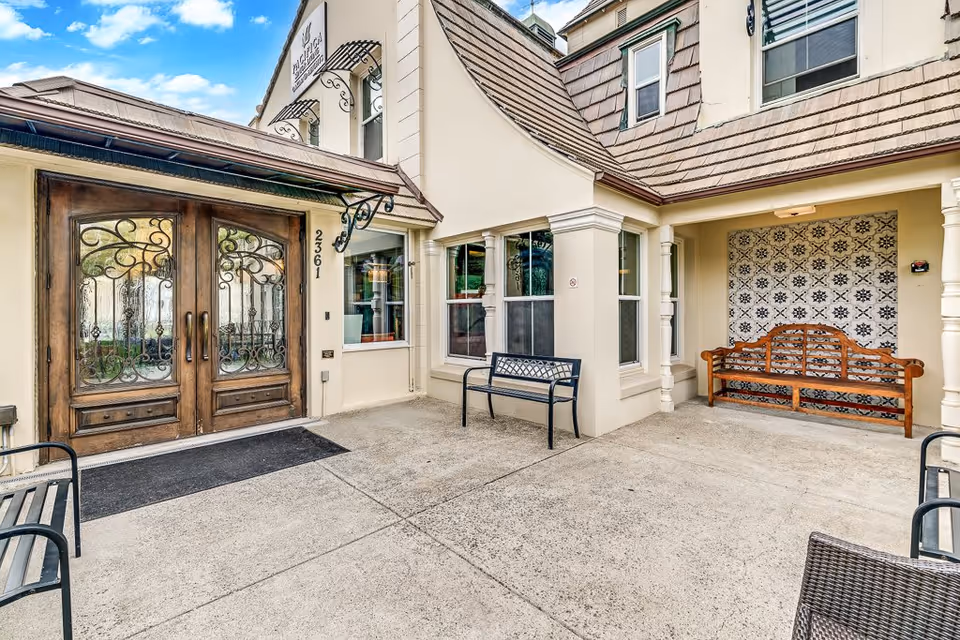 Entrance area of Oakland Heights Senior Living featuring double wooden doors with decorative wrought iron, beige exterior walls, a tiled decorative wall with a wooden bench, and several metal benches on a concrete patio under a partly cloudy sky.