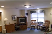 Cozy common room with a television on a wooden cabinet, two rocking chairs by a window, and additional furniture and lamps.
