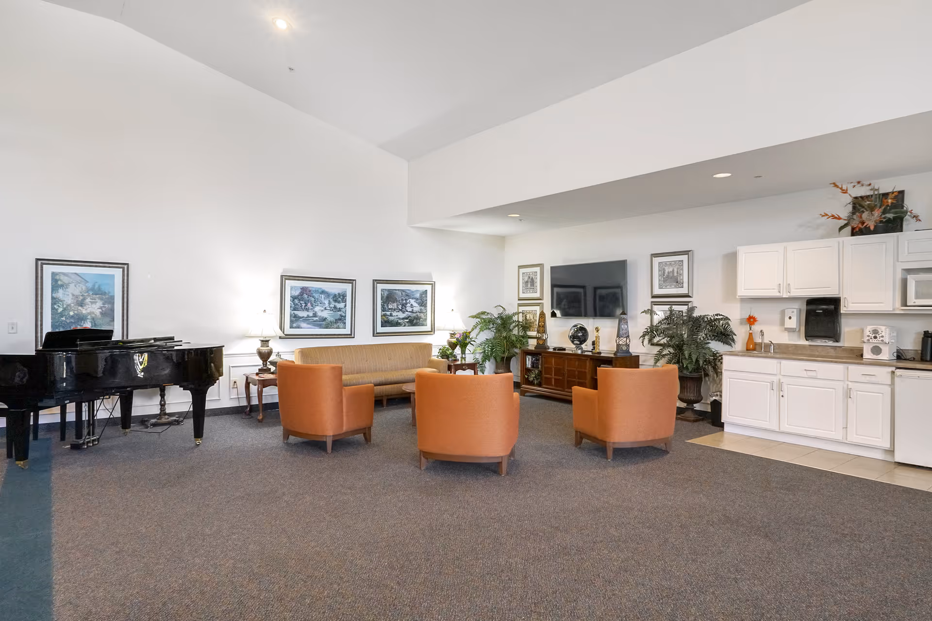 A spacious common area with a grand piano on the left, a beige sofa and three orange armchairs arranged around a coffee table in the center, and a flat-screen TV mounted on the wall above a wooden cabinet. The right side features a kitchenette with white cabinets, a microwave, a sink, and a coffee maker. The room is decorated with framed artwork, table lamps, and potted plants.