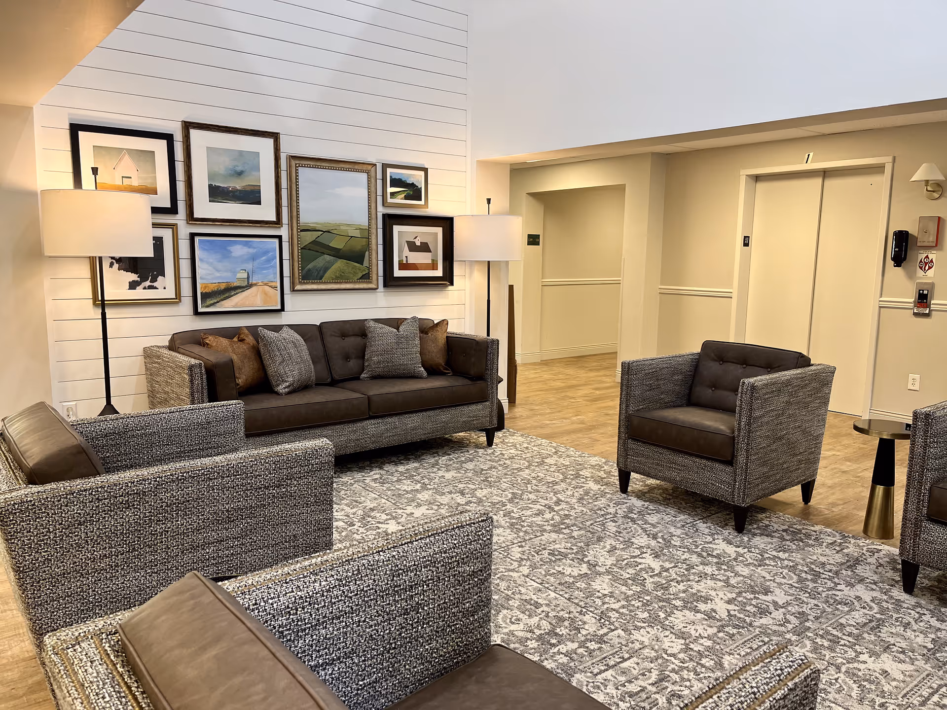 A cozy senior living facility common area with a brown sofa and matching armchairs arranged on a patterned gray and white rug. The walls are light-colored with a gallery of framed landscape and abstract paintings above the sofa. Two floor lamps with white shades provide warm lighting. In the background, there is a hallway and an elevator with closed doors.