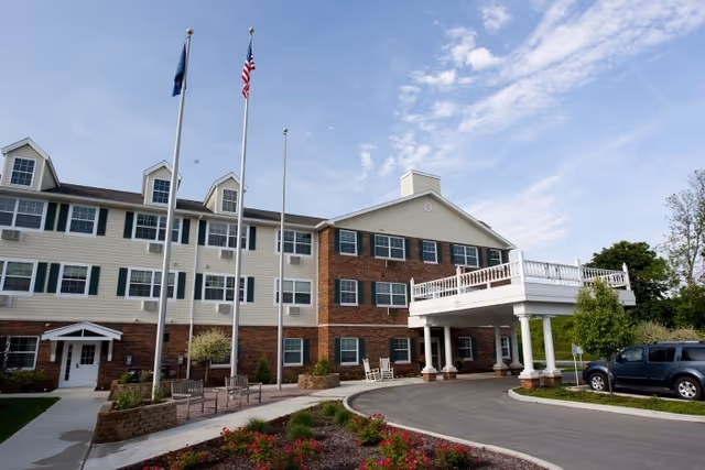 Exterior view of a multi-story senior living facility building with a brick and beige siding facade, multiple windows with green shutters, a covered entrance supported by white columns, three flagpoles with flags, landscaped flower beds, benches, and a parked vehicle on the driveway.