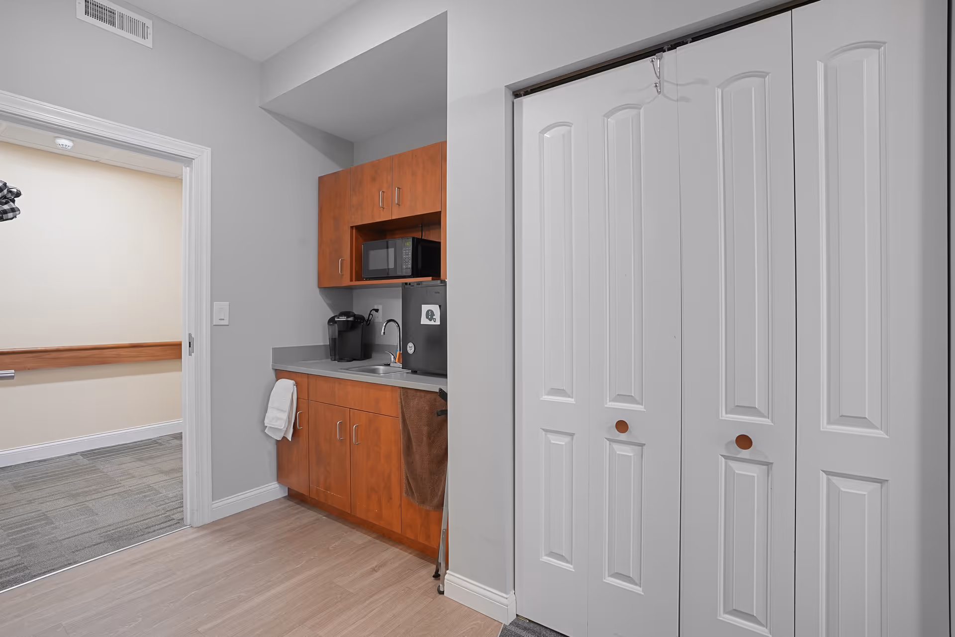 Small kitchenette area with wooden cabinets, a microwave, a mini refrigerator, a coffee maker, and a sink. There are white bi-fold closet doors to the right and an open doorway leading to a hallway with carpeted flooring.