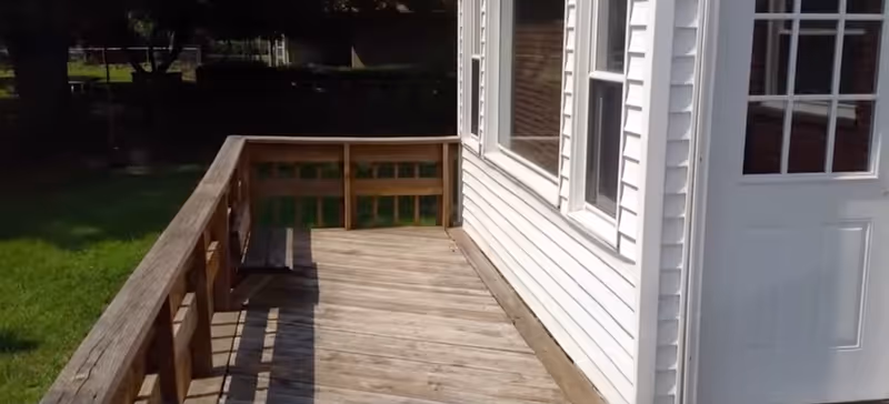 A wooden deck with railing runs alongside a white-sided building with windows and a door overlooking a grassy yard.