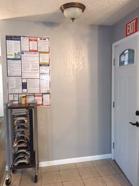 Interior corner of a room with a white door on the right side featuring a small window and an exit sign above it. On the left wall, there is a large employee rights and labor law poster. Below the poster is a metal rack on wheels holding multiple stacks of papers or magazines. The floor is tiled and the walls are painted light gray.