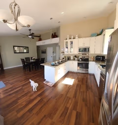 Bright open-plan kitchen and dining area with white cabinets, a central island, stainless steel appliances, hardwood floors, and a small white dog on the floor.