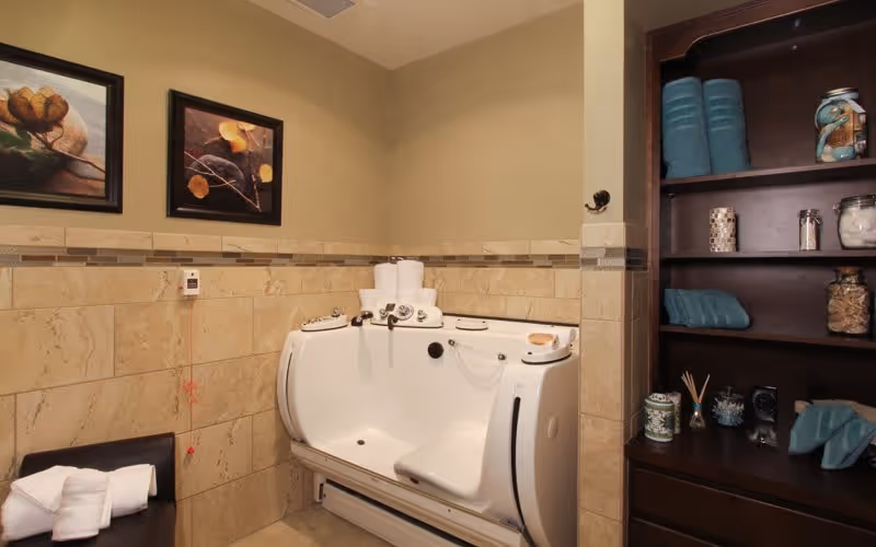 A bathroom with a walk-in bathtub featuring built-in controls and a seat. The walls are tiled halfway up with beige tiles and a decorative border. There are two framed floral paintings on the wall above the bathtub. To the right, there is a dark wooden shelving unit with neatly folded blue towels and various jars and decorative items. A small black chair with folded white towels is positioned to the left of the bathtub.