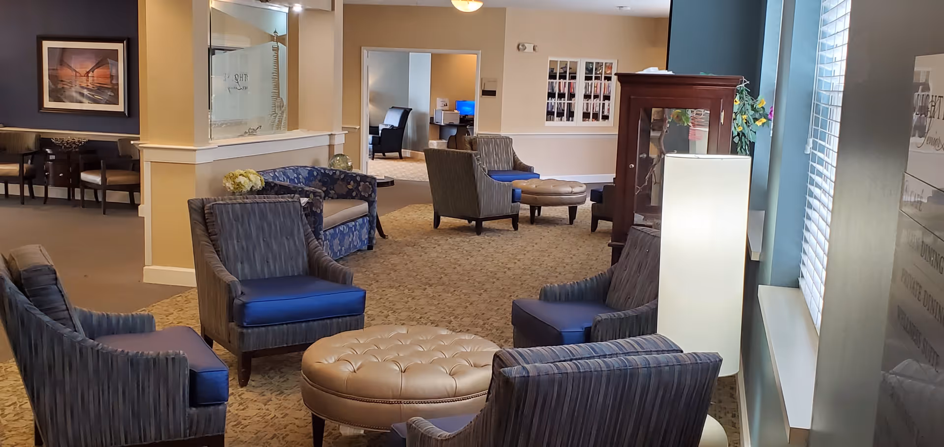 A cozy senior living facility common area with several upholstered armchairs arranged around a round tufted ottoman. The room features carpeted flooring, a tall floor lamp, a wooden display cabinet, and large windows with blinds allowing natural light. In the background, there is a hallway leading to other rooms and a small seating area with a table and chairs.