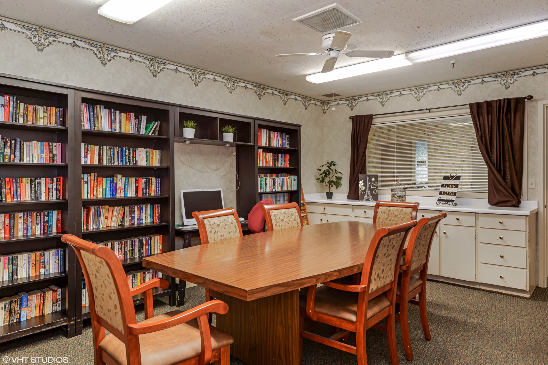 A cozy room with a wooden table surrounded by six chairs with patterned upholstery. Behind the table, there are dark wooden bookshelves filled with books and a computer monitor on a small desk. The room has a ceiling fan, fluorescent lighting, and a window with brown curtains and blinds. Below the window is a white cabinet with multiple drawers and decorative items on top.