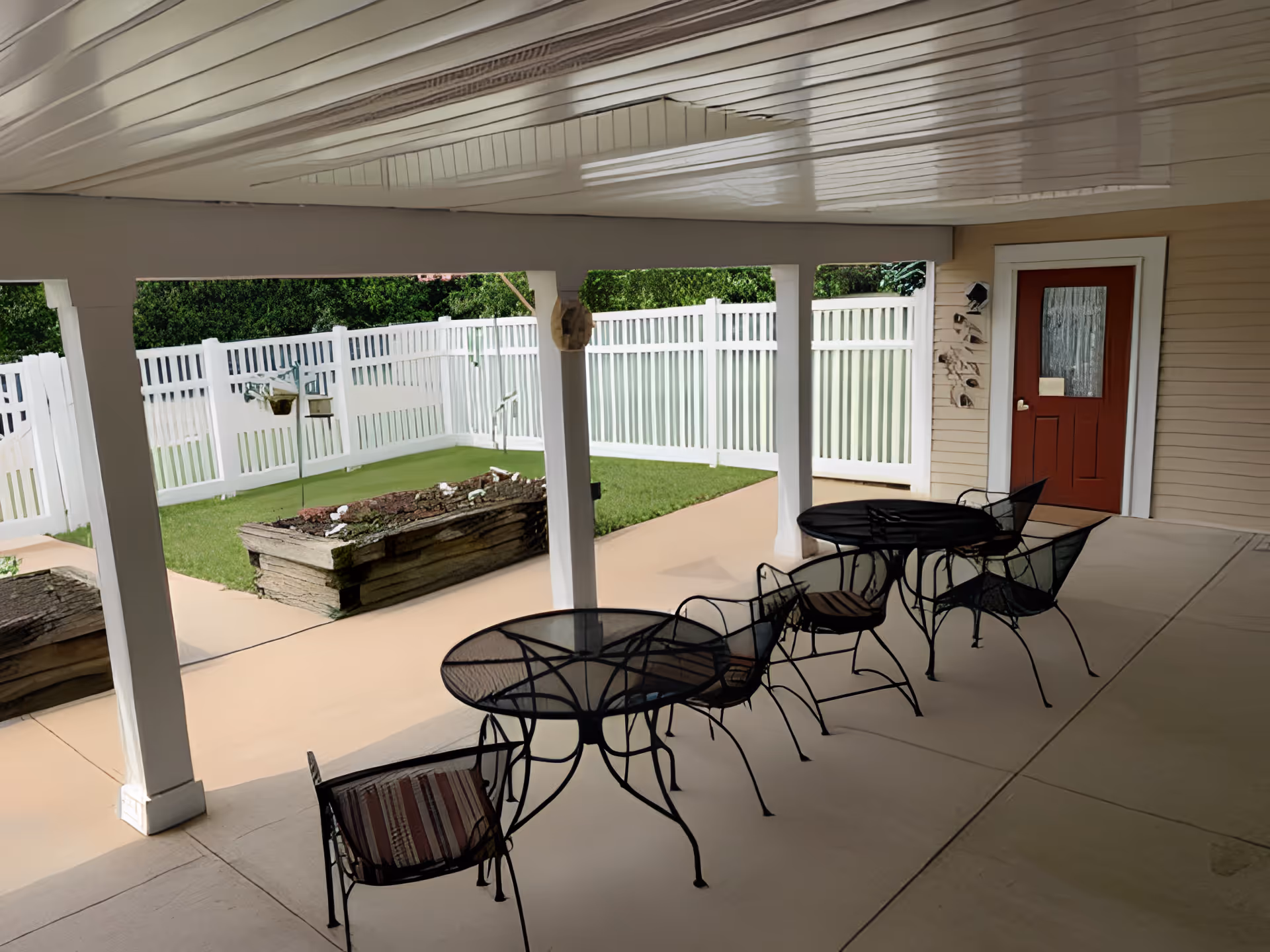 Covered outdoor patio area with metal tables and chairs, overlooking a small fenced garden with raised flower beds and a white picket fence. A red door is visible on the beige siding of the building.