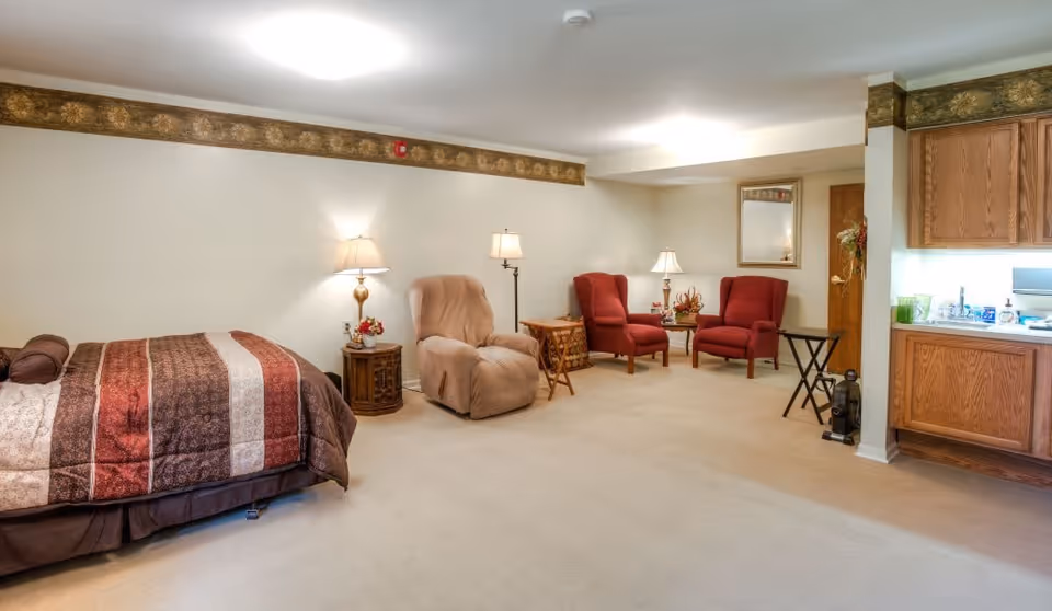 A spacious room with a bed on the left side covered with a brown and red striped comforter. Next to the bed is a wooden nightstand with a lamp and a small flower arrangement. In the middle of the room is a beige recliner chair with a floor lamp behind it. Towards the back wall, there are two red armchairs with a small wooden table between them, topped with a lamp and decorative items. On the right side of the room is a kitchenette with wooden cabinets, a sink, and various kitchen items. The walls are light-colored with a decorative border near the ceiling, and the floor is carpeted in a light beige color.