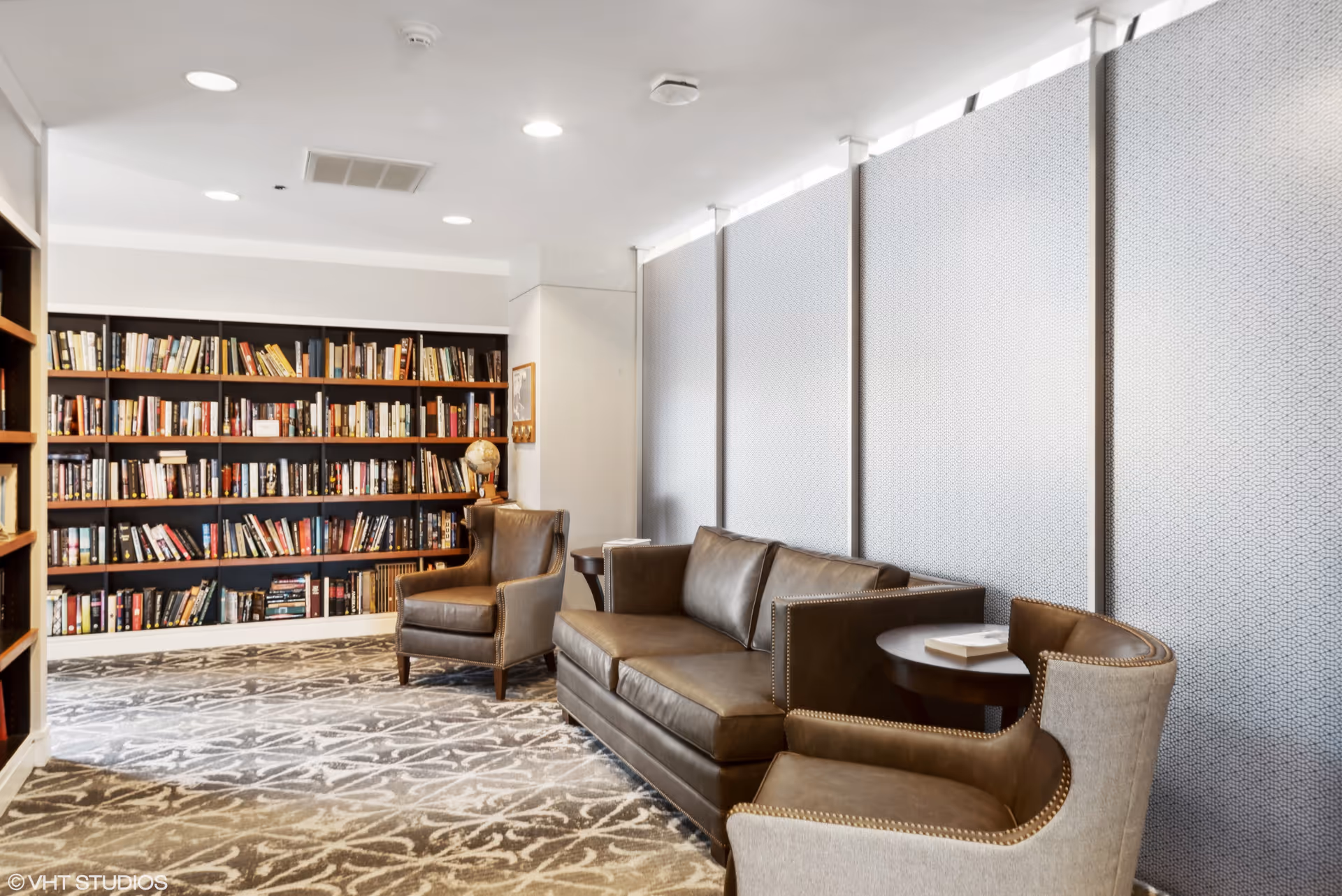 A cozy reading lounge area with a large bookshelf filled with books, a brown leather sofa, two brown leather armchairs, and a small round wooden side table with a book on it. The room has patterned carpet flooring and light-colored walls with tall windows covered by patterned blinds.