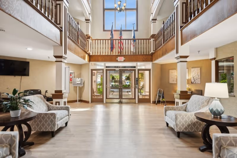 Spacious and well-lit lobby area of a senior living facility with two patterned armchairs and side tables on each side, a large window above the entrance doors letting in natural light, and flags displayed on the balcony railing above the entrance.
