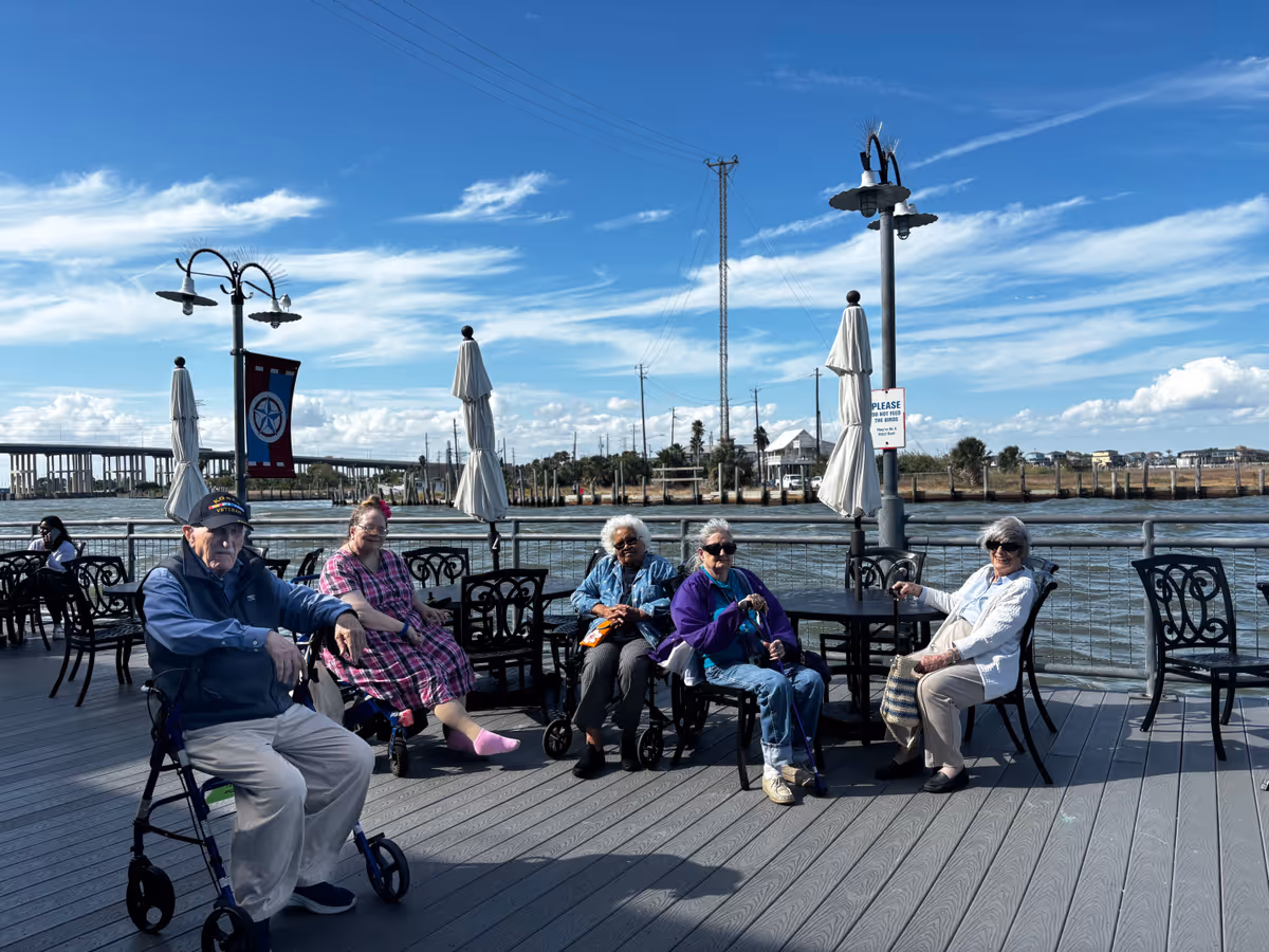 Five elderly people sitting on chairs and wheelchairs on a wooden deck by the water under a blue sky with scattered clouds. There are tables, umbrellas, and lamp posts around them, with a bridge and buildings visible in the background.