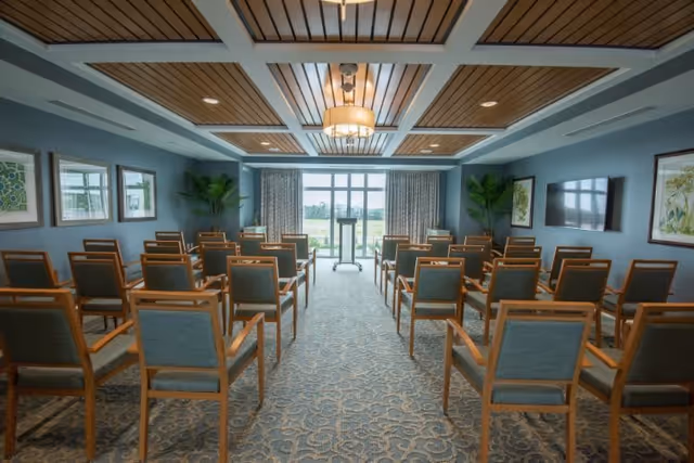 A conference or meeting room with rows of wooden chairs with blue cushions arranged facing a podium and large window with curtains. The room has blue walls, framed artwork, potted plants, a wall-mounted TV, and a ceiling with wooden panels and recessed lighting.