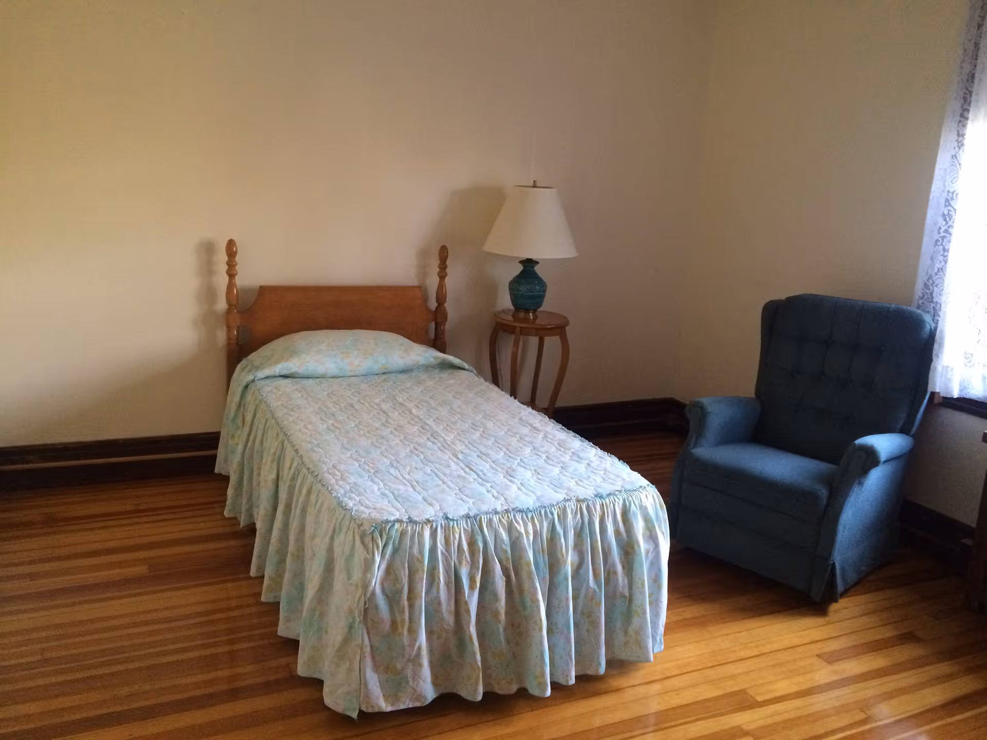 A simple bedroom with a single wooden bed covered with a light blue floral bedspread, a small wooden side table with a green lamp, and a blue upholstered armchair near a window with white lace curtains. The room has wooden floors and beige walls.