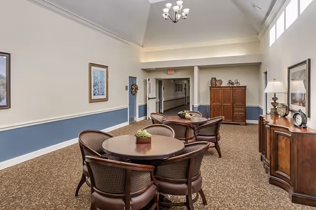 Interior view of a senior living facility common area with a round wooden table surrounded by four chairs in the foreground, another similar seating arrangement further back, beige walls with blue wainscoting, carpeted floor, framed artwork on the walls, and a wooden cabinet with decorative items and a lamp on the right side.