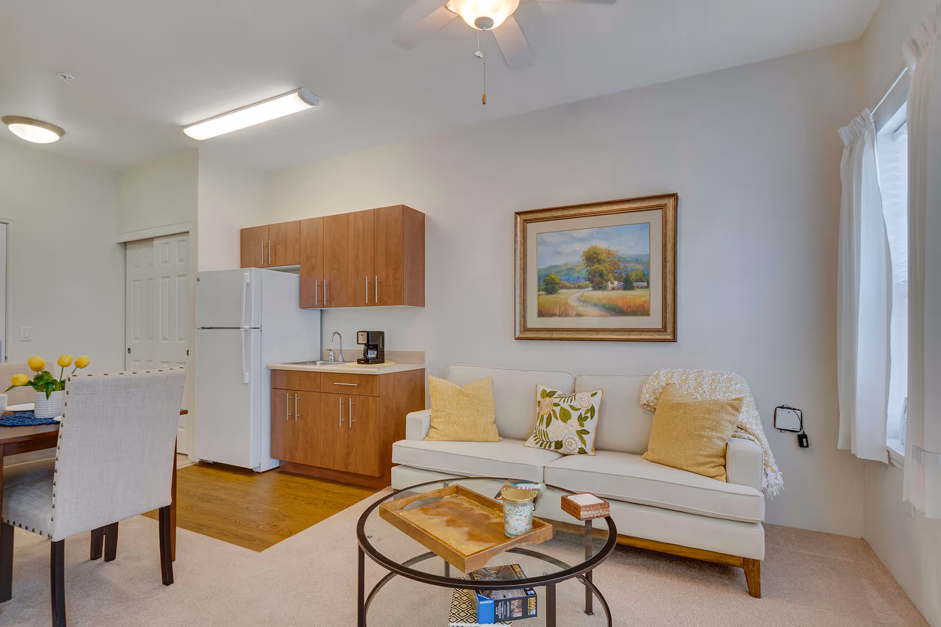A cozy living area with a beige sofa adorned with three decorative pillows and a throw blanket. Above the sofa hangs a framed landscape painting. In front of the sofa is a round glass coffee table with a wooden tray and some small items on it. To the left, there is a small kitchenette with wooden cabinets, a white refrigerator, a sink, and a coffee maker. A dining table with upholstered chairs and a vase of yellow flowers is partially visible. The room has light-colored walls, a ceiling fan, and a window with white curtains allowing natural light to enter.