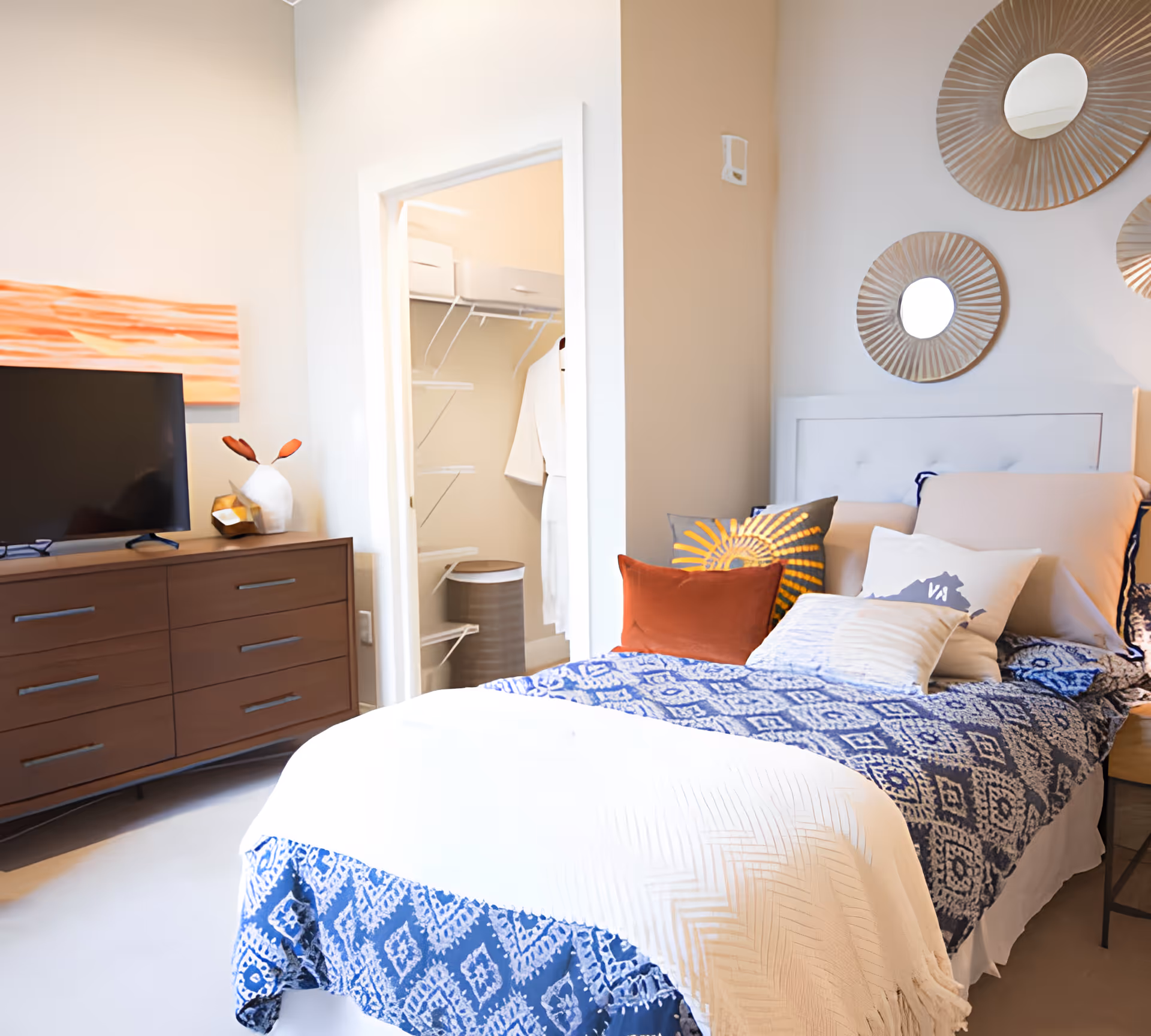 A cozy bedroom with a bed covered in a blue and white patterned blanket and multiple pillows, including one with a sun design and another with a map of Virginia. There is a wooden dresser with a TV on top and decorative items beside it. The room has a doorway leading to a walk-in closet with shelves and hanging clothes. Three round decorative mirrors are mounted on the wall above the bed.