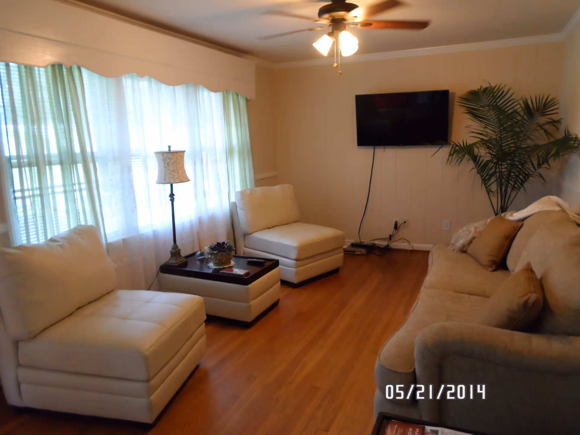 Cozy living room with a beige sofa, two white armless chairs, a coffee table, wall-mounted TV and large curtained window.